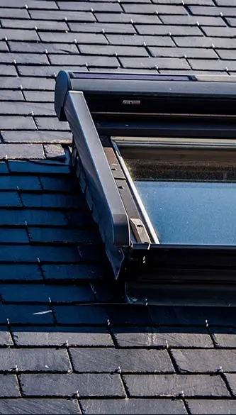 Une fenêtre de toit, type Velux, est installée sur une toiture en ardoise sombre. Les tuiles d'ardoise sont disposées avec précision selon la technique de l'alignement à joints croisés, garantissant une couverture étanche et uniforme.