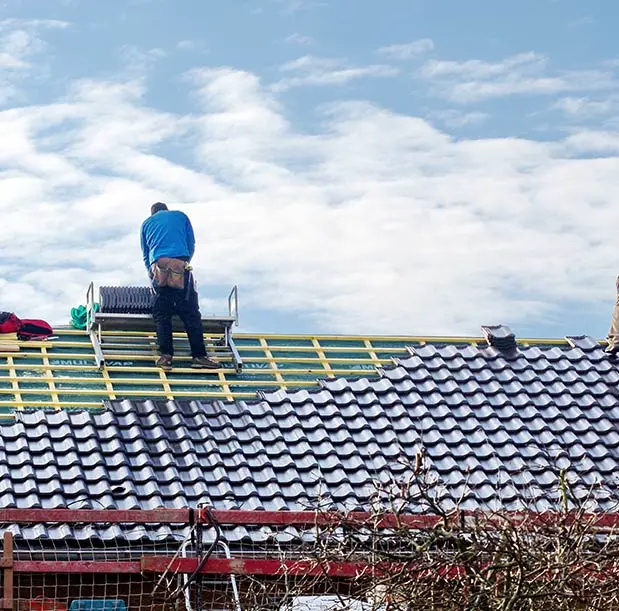 Un artisan couvreur travaille sur une toiture en tuiles grises, posées sur une structure en bois. Une échelle de toit est utilisée pour faciliter l'accès aux différentes sections de la toiture