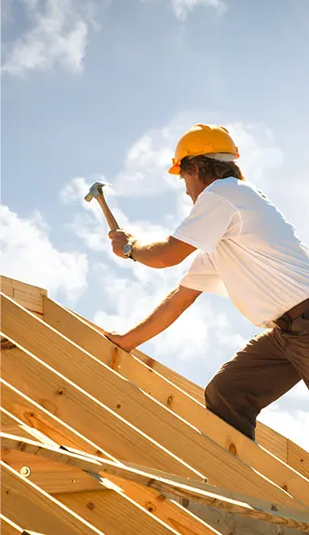 Un charpentier, portant un casque de sécurité jaune, travaille sur une charpente en bois. Il utilise un marteau pour fixer les éléments de la structure