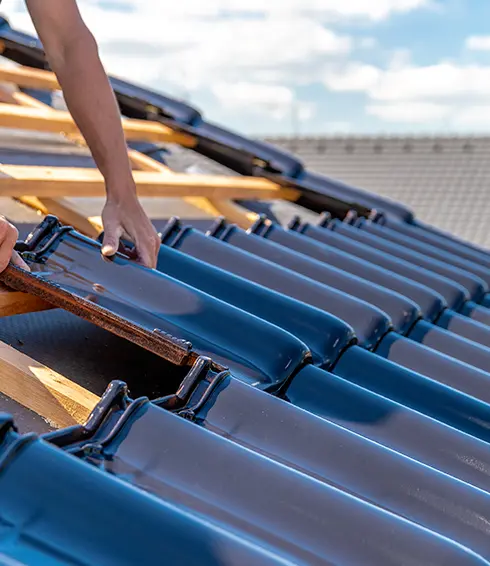 Deux artisans couvreurs installent des tuiles bleues sur une charpente en bois. L'un ajuste les tuiles avec précision, tandis que l'autre, équipé d'un casque et d'un harnais de sécurité, travaille sur une autre section du toit