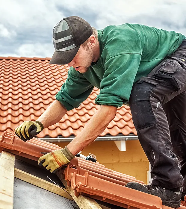 Un artisan couvreur installe des tuiles en terre cuite sur une toiture. Vêtu d'un pull vert et d'une casquette, il travaille minutieusement, ajustant les tuiles pour assurer un alignement précis. Les tuiles sont fixées sur une structure en bois