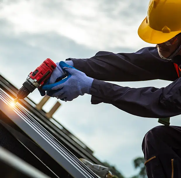 Un artisan couvreur zinguer portant un casque de sécurité jaune et des gants bleus utilise une visseuse électrique pour fixer des panneaux métalliques sur une toiture. Il travaille avec précision pour assurer la solidité et l'étanchéité de la couverture, garantissant ainsi une installation durable et sécurisée